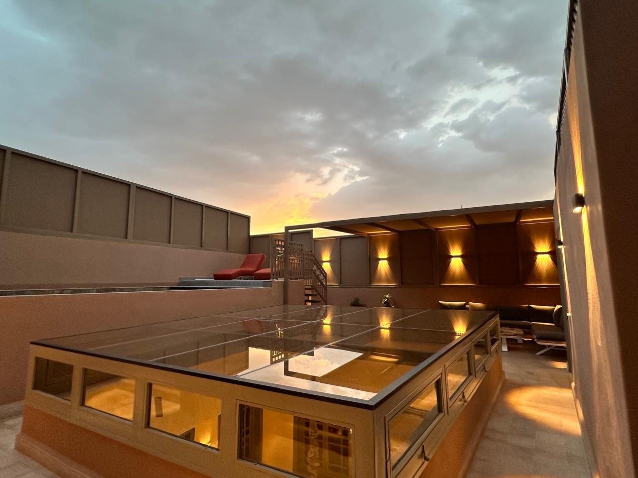 The rooftop terrace at sunset, wall lights glowing above the lounge and the glazed roof of the patio below