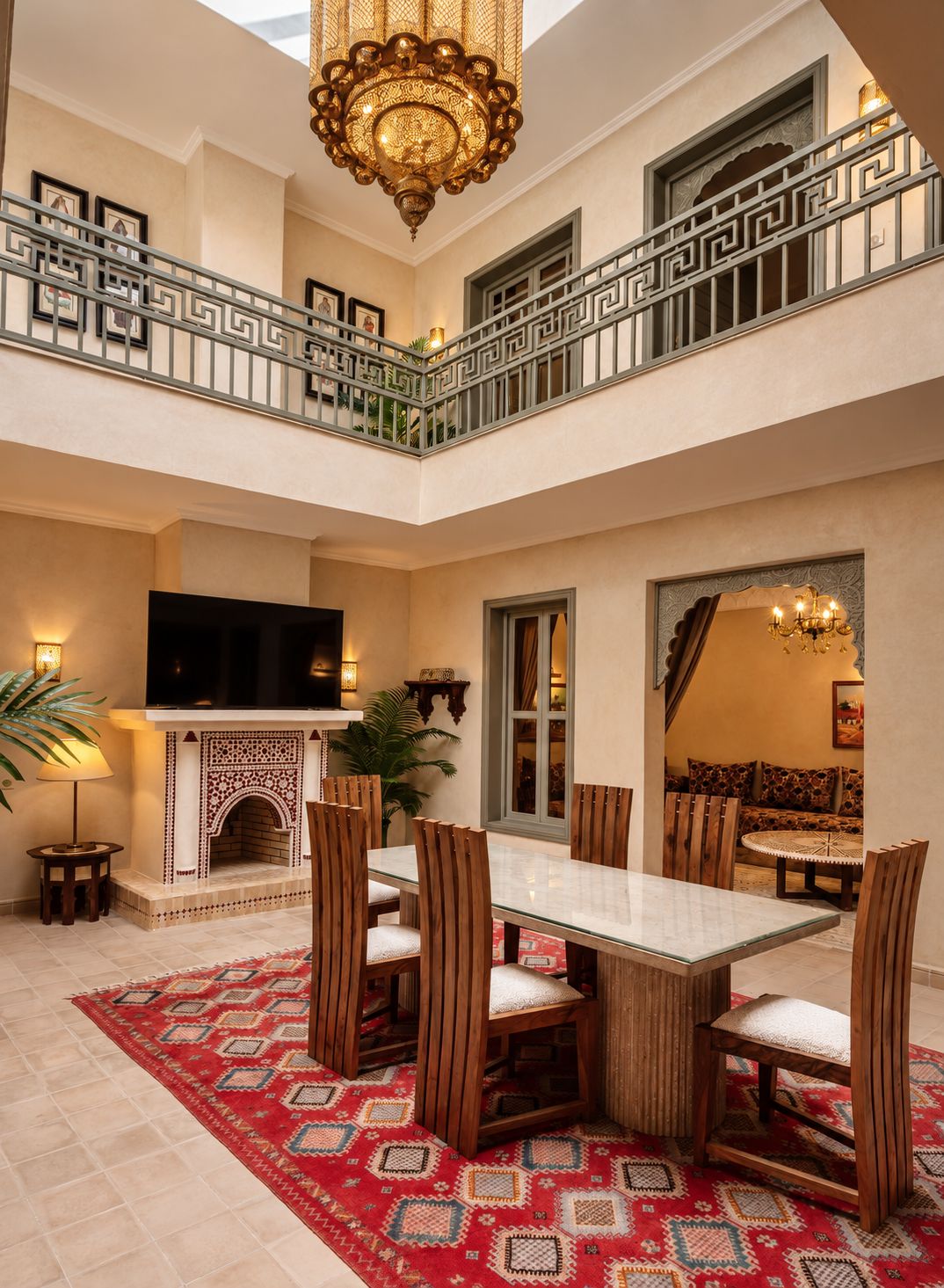 The patio: a zellige fireplace and dining table beneath the upper gallery, the brass chandelier hanging through two floors, the Moroccan salon through an archway
